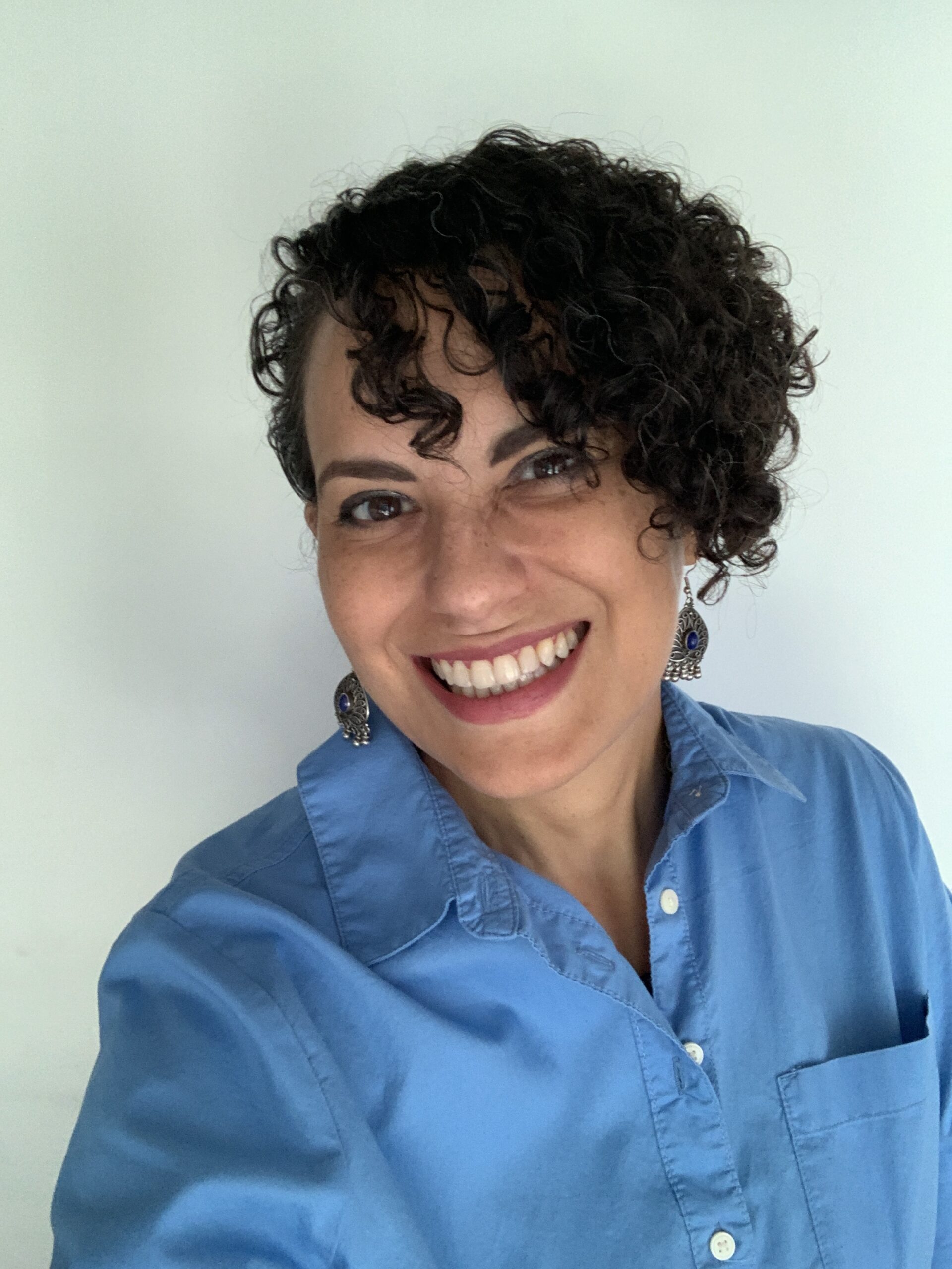 Elisa Burgos-Ojeda, a smiling Latina with short dark curly hair, silver earrings, and a blue shirtdress.
